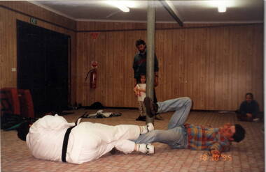 Two boys are lying on the floor during a judo bout while an adult and little child and another boy looking on.
