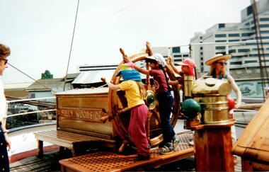 Three children are turning a wheel on a ship's deck with two adults looking on. The city skyline is in the middle distance.