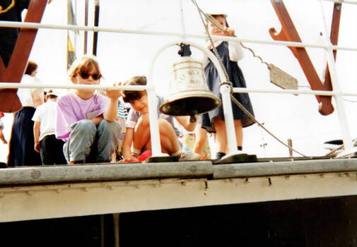 Three little girls are crouched down on the deck of a ship and looking at the bell.