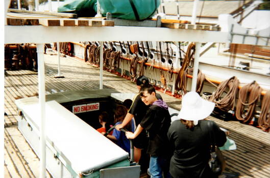 Five children with an adult are going below decks on a ship. A lot of the ship's rigging is visible on the RHS of the image.