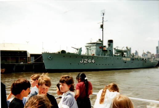 A view of a naval vessel docked and a group of children on a ferry looking at it.
