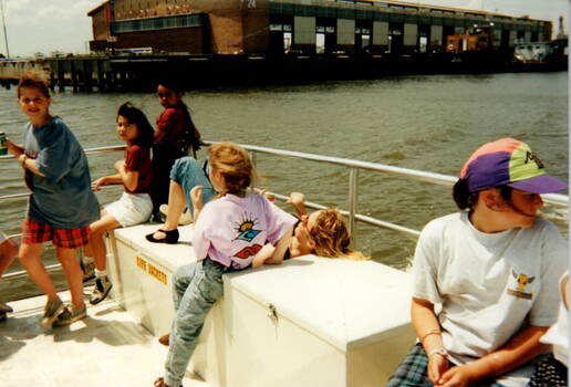 Six children are sitting on the deck of a ferry.A large cargo storage shed is in the background.
