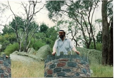 A coloured photograph of a man standing at the top of a castle turret in a woodland setting.