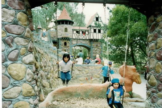 Two girls are on swings in the foreground with a group looking at a tudor style arch in the background in an amusement park.