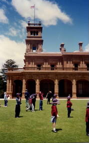 A group of children lined up on an expanse of lawn outside a grand Italianite mansion. They are getting ready to play some games.