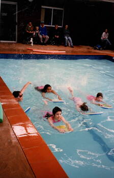 Five children are swimming in an indoor pool with the aid of kick boards while some adults are seated at the side watching.