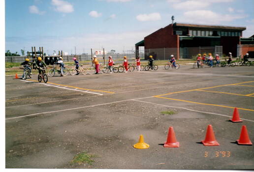 A coloured photograph of a lot of children partaking in a lesson on bike riding on an large asphalted open space. A sports pavilion is in the background.
