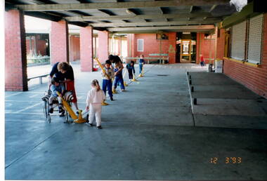 A coloured photograph of five young children being supervised by two adults in a sheltered courtyard. The children are playing with a softball bats and balls. 