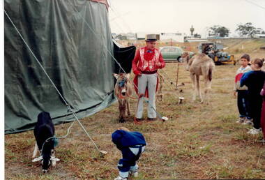 A coloured photograph of a circus ringmaster, who is holding a pony and talking to them about the donkey, the camel and the goat that are some of the circus animals.There are vehicles in the background and an air control tower is visible on the skyline.