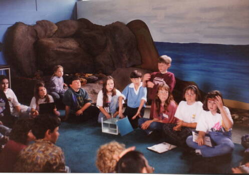 A group of children seated on a floor listening to a person giving a talk. Some of them have notebooks and writing materials to take notes.