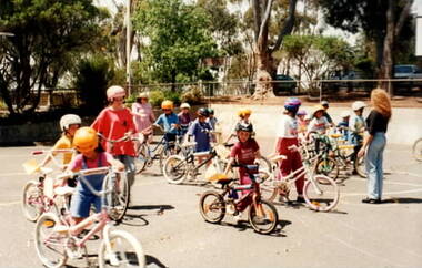 A group of children with their bicycles standing in an asphalted area and listening to an adult talking to them during a bicycle education class.