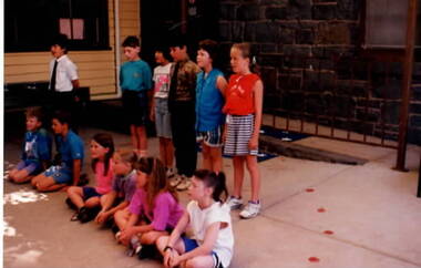 Five boys and seven girls are standing and sitting in an open area and performing for an audience.