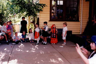 A group of children with an adult are gathered around in a covered-in outdoor area with four of the children are dressed in traditional costumes and lined up in front of the others.