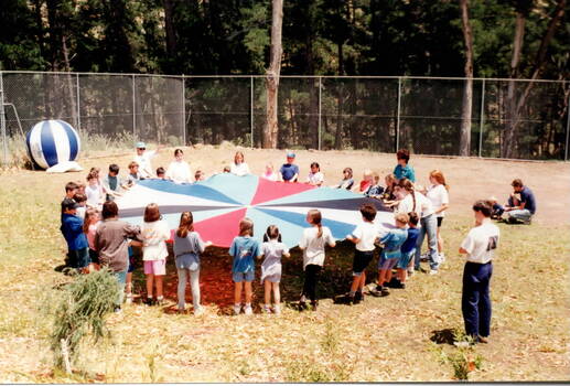 A group of children are standing in a grassed area, in a large circle while holding onto a large coloured circular banner with an adult supervising the group.