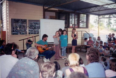 Four children are singing to the accompaniment of a man playing a guitar, in front of an audience made up of adults and children,
