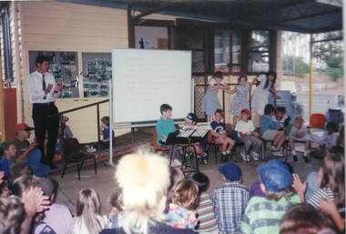 Eight children are performing on a variety of musical instruments including keyboard, recorders and percussion in front of an audience. A teacher is standing to the left of the group observing them.