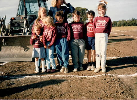 A lady standing behind seven students from Bulla Primary School in an open area and in fro0nt of a large grader.