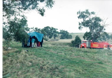 Children huddled under a large waterproof sheet to shelter from a sudden shower of rain with the park ranger's truck which they were all on is parked a short distance from them.