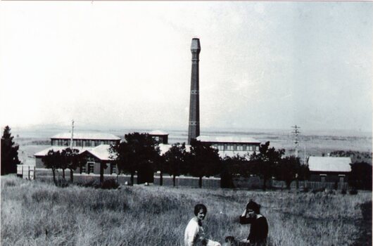 A large brick building with a tall chimney overlooking open rural countryside with two women sitting on the grass in the foreground. 