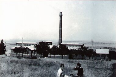 A large brick building with a tall chimney overlooking open rural countryside with two women sitting on the grass in the foreground. 