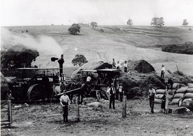 Eleven men are gathering hay with the aid of a steam traction engine in a wide open valley. A hillside in the distance has been terraced and there is a house on the hill in the distance.