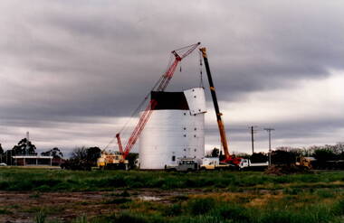Two trucks and a crane are dismantling a metal wheat silo, which is standing in an open area. A CFA pavilion is visible in the distance.
