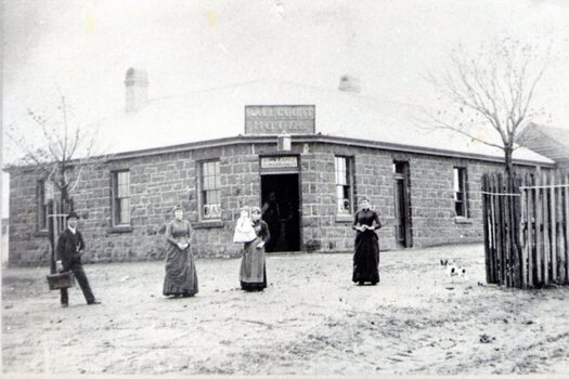 A man and three ladies are standing outside a single story blue stone hotel building known as the Ball Court Hotel. IT has a corner entrance.