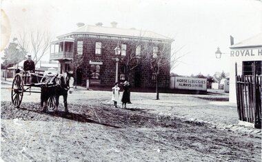 A street photograph featuring an intersection with a two storey bluestone building on one corner and a partial view of a single storey rendered building on the opposite corner. Two children are standing on the side of the road and a man is in a horse-drawn jinker.