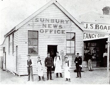 A photograph of a weatherboard news office building with six men standing the front. Three of the men are wearing white aprons.