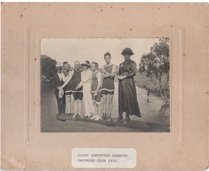 Seven people are standing on a diving board that is over a natural water hole. Four men are dressed in swim wear and two ladies are in full length dresses. A man standing on the end of the board is wearing a suit.