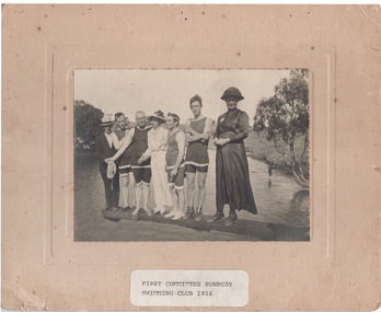 Seven people are standing on a diving board that is over a natural water hole. Four men are dressed in swim wear and two ladies are in full length dresses. A man standing on the end of the board is wearing a suit.