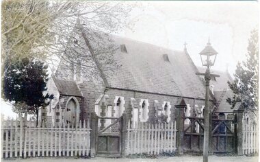A photograph of a small bluestone church with a slate roof with a wooden picket fence and two gates across the front. A gas lamp is on the pathway.