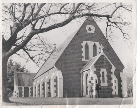 A black and white photograph of a blue stone church with white rendered columns around the windows and door and a pitched slate roof. Two men are standing beside the main entrance.  