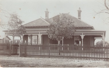 A post card of a sepia photograph of an Edwardian style brick house with a tiled roof and a return verandah. The house has a picket front fence.