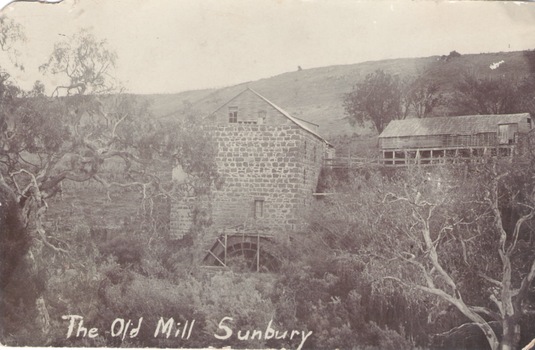 A photograph of a an old bluestone mill building sited on the bank of a watercourse. he remains of the water wheel are at the rear of the building.