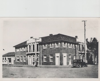 A two storey brick hotel building sited on a corner with a black sedan car parked near the corner.