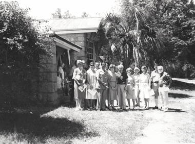 Nineteen people are standing in a garden outside a homestead, which is constructed from stone with a galvanised roof.