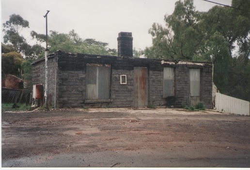  A photograph of a damaged blue stone building. There is no roof and the windows and doors are covered in with sheets of corrugated iron. 