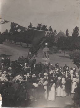 A crowd of people are gathered around a monument for a ceremony. Flags are festooned above the crowd and an open parkland is in the background as well as a church.