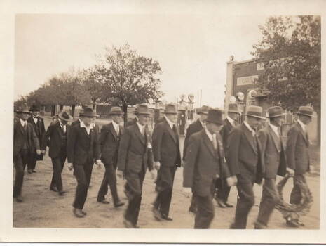 Men dressed in suits are marching in file along a street. One man holds a wreath to place on a memorial.