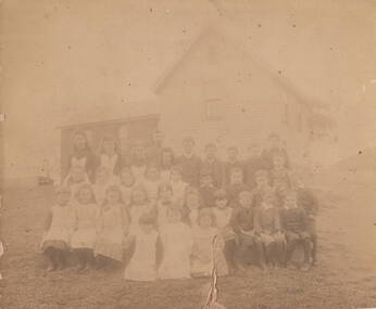  An old photograph of children seated in front of a small weatherboard school, their teacher is standing behind the pupils.