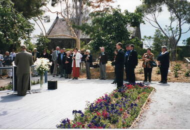 A man is standing in front of a lectern and addressing a group of people. Small church is in the background.