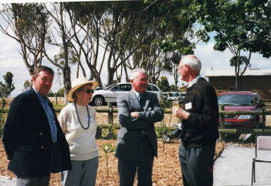 A lady and three men are conversing while standing in a garden. 