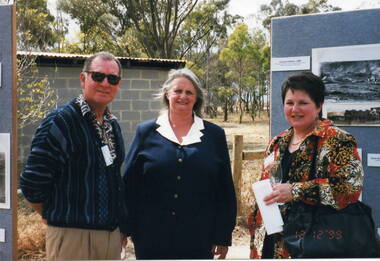 Two Ladies and a man are standing in a garden at an official function.