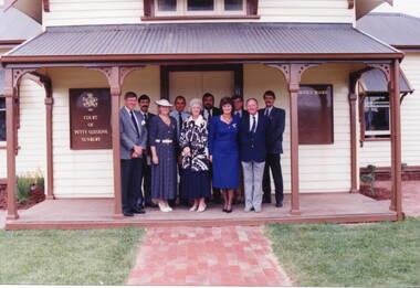 Nine people at an official function are standing on the verandah of a weatherboard building. 