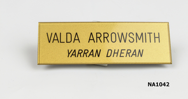 Gold coloured oblong badge with 'Valda Arrowsmith/Yarran Dheran' printed in black.