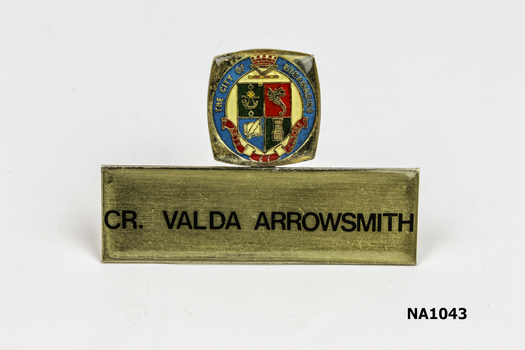 Square 'City of Nunawading' and coat of arms sitting of top of gold coloured oblong badge with 'Cr. Valda Arrowsmith' . Printed in black.
