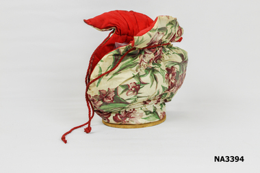 Padded tea cosy made up of floral plastic cover with red cotton lining fixed to oval plywood base. 