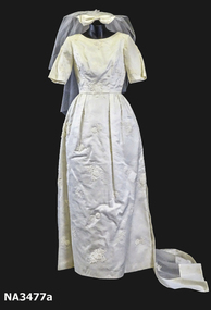 1963 Wedding dress of cream satin; floor length, short sleeves, round neckline, fitted at waist.