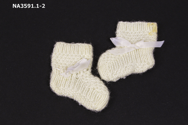 A pair of creamy white knitted wool booties, 1964, with nylon ribbon threaded around ankles. 